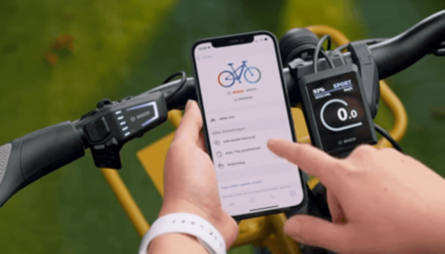 Bild eines Telefons, auf dem die Bosch E-Bike Flow App läuft. Mit einem Elektrofahrrad mit Telefonhalterung im Hintergrund.
